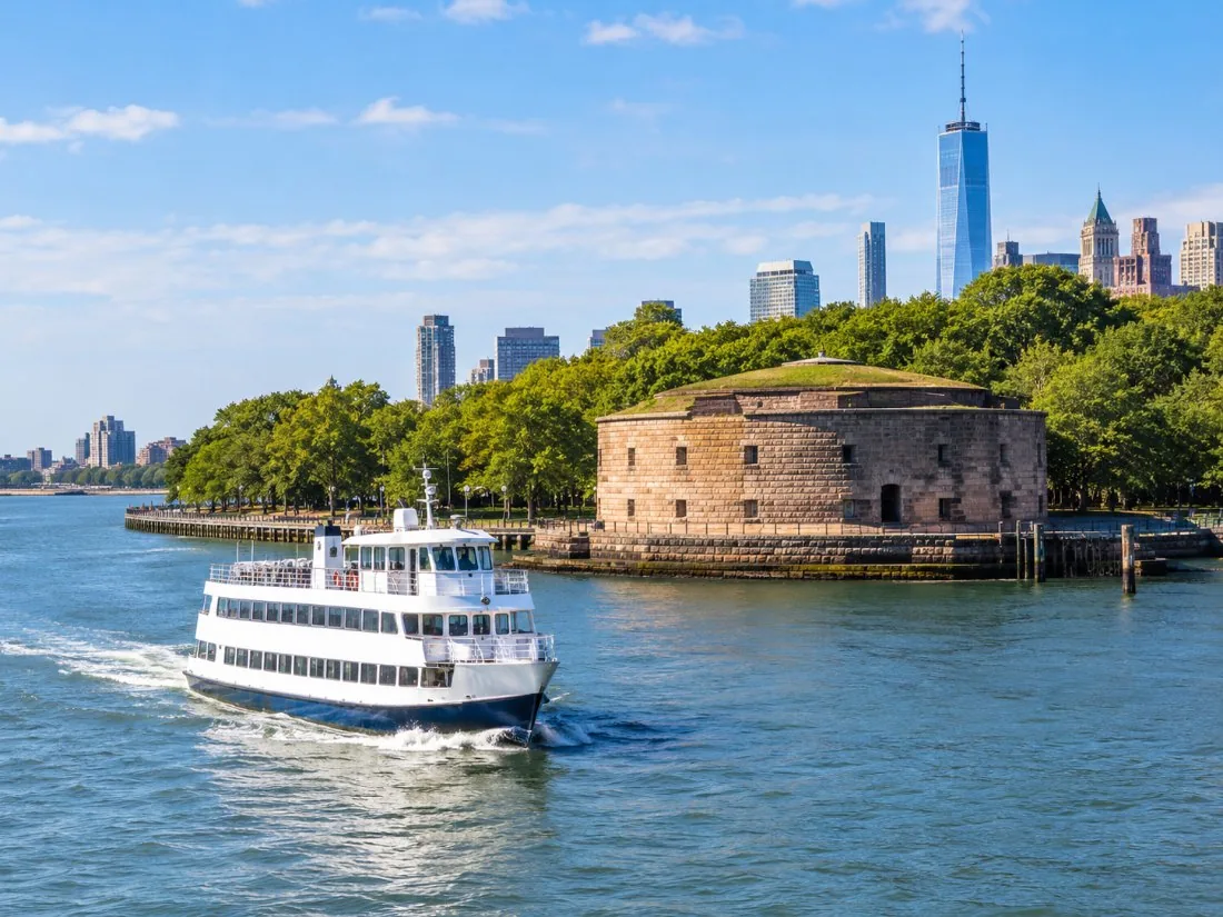 Un ferry blanco de dos cubiertas alejándose de un fuerte de piedra en la punta de Manhattan en una mañana soleada