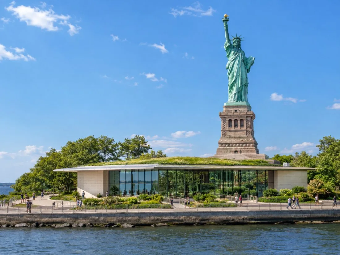 Un pabellón de museo bajo de vidrio y granito en Liberty Island, con la estatua detrás